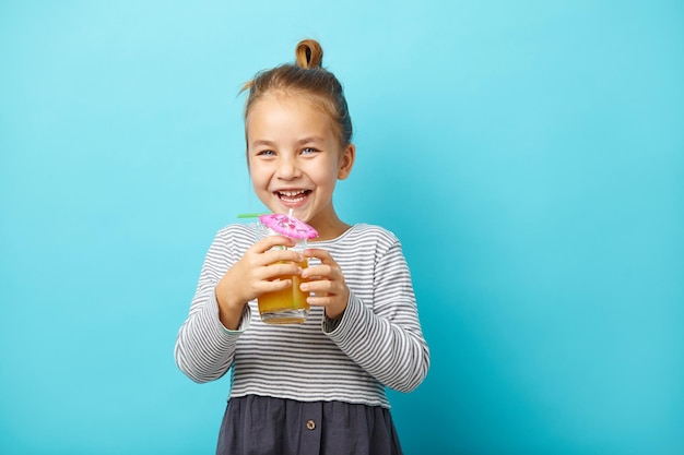 Cheerful little girl drinks orange juice and laughs stands on blue isolated background Studio portrait of happy child