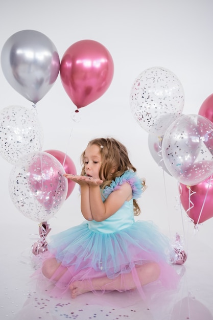 Cheerful little girl in a festive dress with a tutu skirt plays with confetti on a white background with balloons