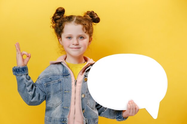 Cheerful little girl holding speech bubble and doing ok sign on yellow background