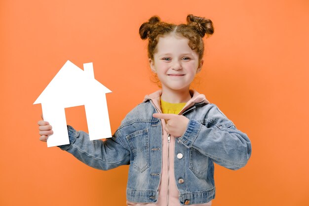 Cheerful little girl holding a white cardboard house and pointing at it on orange background