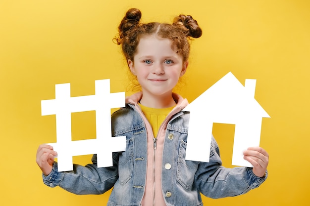 Cheerful little girl holding a white hashtag and house on yellow background