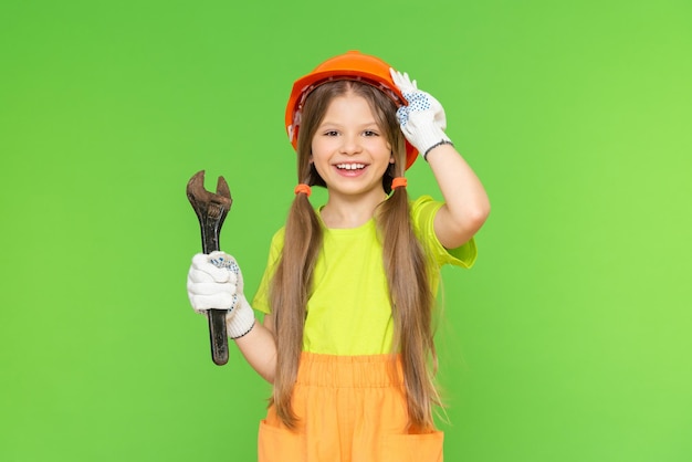 A cheerful little girl holds a construction safety helmet and a wrench. Construction and repair. Green isolated background. Copy space.