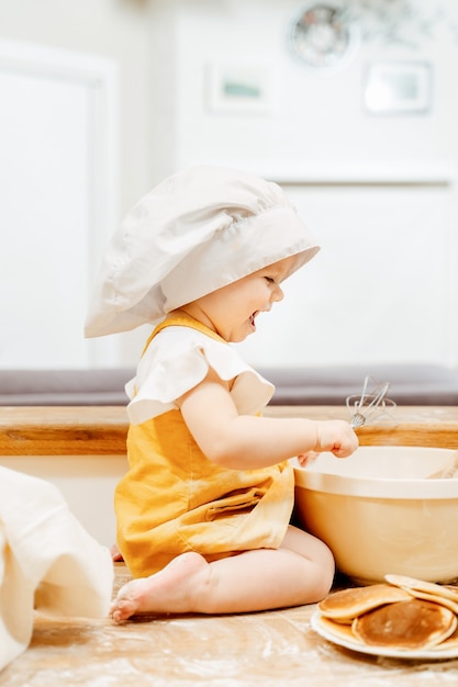 白い帽子をかぶった陽気な少女は、テーブルの上に座っている小麦粉のプレートに泡立て器で生地を準備します