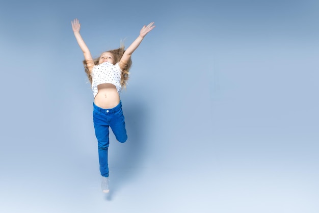 점프하는 쾌활한 어린 소녀가 스튜디오의 파란색 배경에서 손을 들어 올립니다