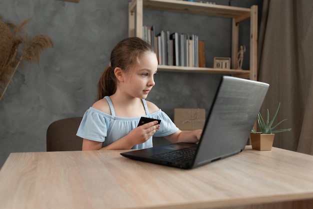 cheerful little girl makes online purchases using a laptop and a credit card while relaxing at the table at home Copy space