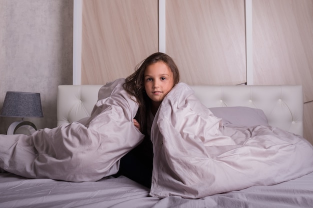 A cheerful little girl in pajamas is sitting under the blanket in bed