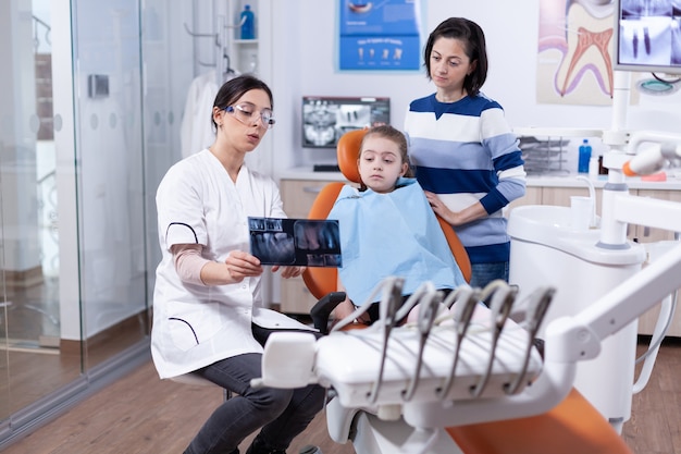 Cheerful little girl sitting in dental office while doctor showing something on radiography. Stomatologist explaining teeth diagnosis to mother of child in health clinic holding x-ray.