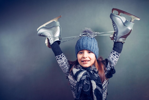 Cheerful little girl in warm sweater and hat holding figure skates.