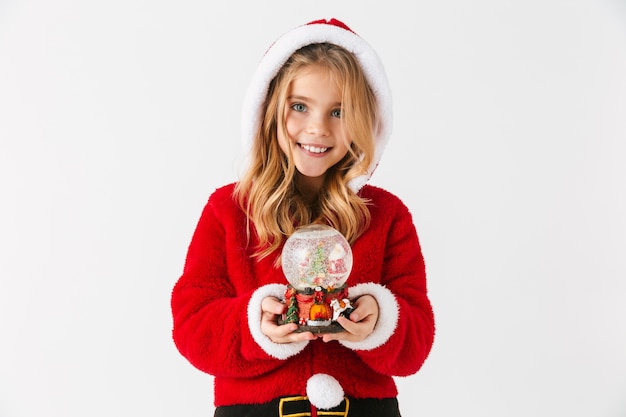 Cheerful little girl wearing Christmas costume sitting isolated, holding a snow globe