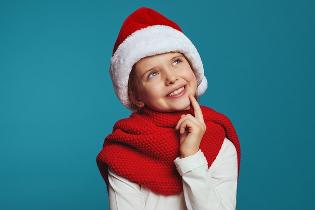 Cheerful little girl wearing christmas hat and red scarf looking at camera