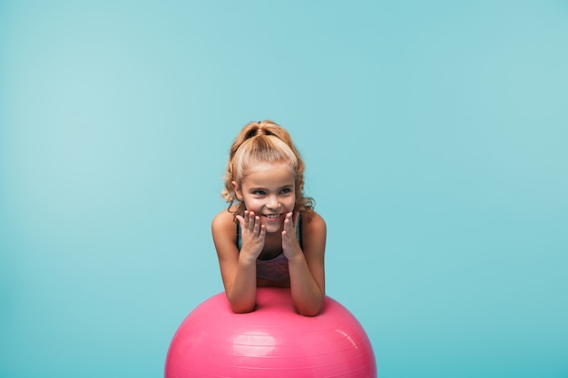 Cheerful little girl wearing sport clothes leaning on a fitness ball isolated over blue wall