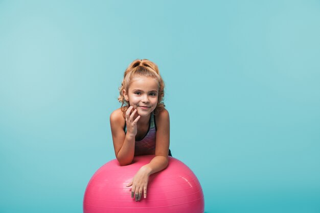 Bambina allegra che indossa abiti sportivi che si appoggia su una palla fitness isolata sopra la parete blu