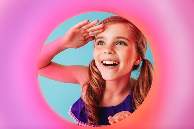 Cheerful little girl wearing swimsuit standing isolated over blue wall, playing with inflatable ring