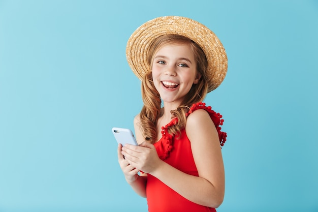 Cheerful little girl wearing swimsuit and summer hat standing isolated over blue wall, using mobile phone