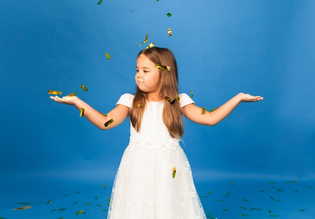 Cheerful little girl in white dress dancing on confetti isolated on blue studio background.