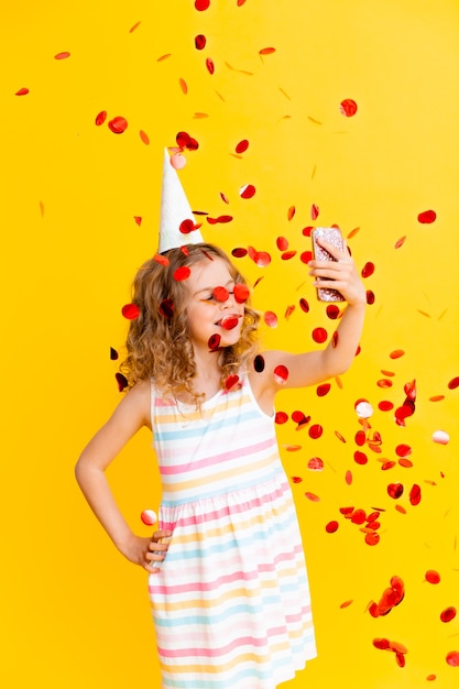 Cheerful little girl with blond curly hair is celebrating her birthday. The child holds the phone, takes a selfie in the rain of confetti. Close-up portrait on yellow background.