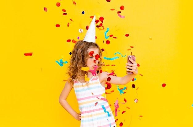 Cheerful little girl with blond curly hair is celebrating her birthday. The child holds the phone, takes a selfie in the rain of confetti. Close-up portrait on yellow background.