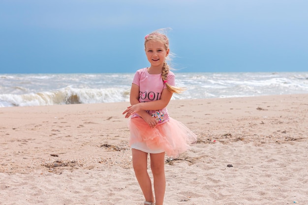 Cheerful little girl with long blonde hair in pink tulle skirt walking empty sea beach alone Beautiful little princess