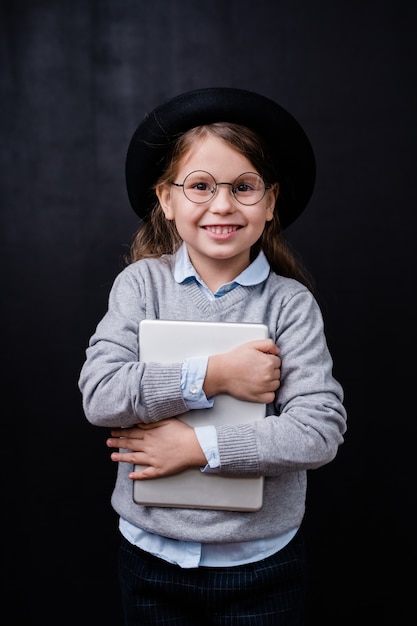 Cheerful little girl with toothy smile holding digital tablet by chest while standing in isolation against black space