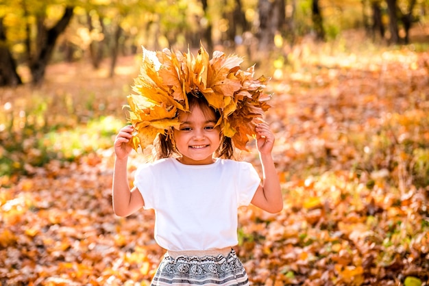 Cheerful little girl with a wreath of maple leaves on her head in a white tshirt on an autumn day