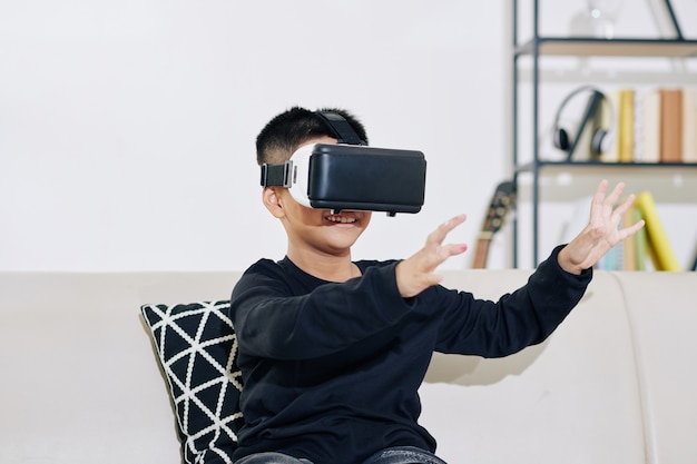 Cheerful little kid in virtual reality headset playing game at home