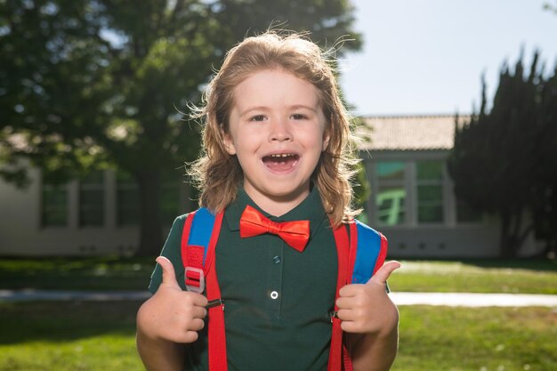 Cheerful little school boy in school uniform with big backpack standing near school Back to School Kids preschool education concept