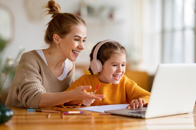 Photo cheerful little schoolgirl in headphones talking to the teacher while studying remotely via laptop at home with happy mom sitting nearby and giving support