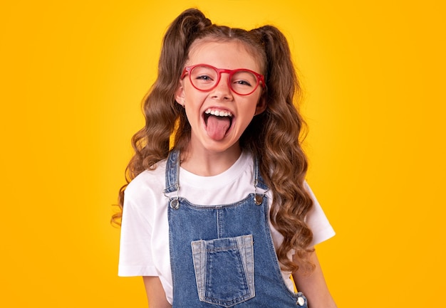 Cheerful little schoolgirl with curly ponytails wearing trendy denim overall and eyeglasses having fun and showing tongue