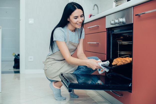 Cheerful long haired lady squatting down near the open oven and taking a baking pan with croissants out of it