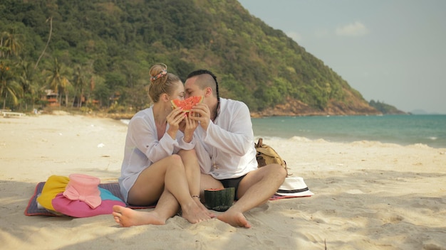 The cheerful love couple holding and eating slices of\
watermelon on tropical sand beach sea. romantic lovers two people\
spend summer weekend.