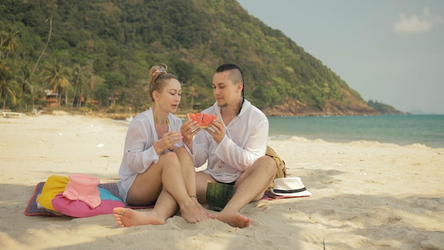 The cheerful love couple holding and eating slices of watermelon on tropical sand beach sea