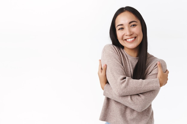 Ragazza asiatica adorabile adorabile allegra, studentessa in maglione accogliente, abbracciandosi, abbracciando il proprio corpo esprime l'amore per se stessi e l'accettazione, sorridendo ridendo gioiosa