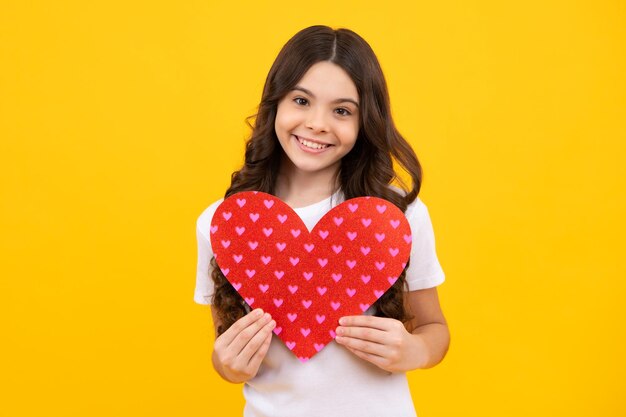 Cheerful lovely romantic teen girl hold red heart symbol of\
love for valentines day isolated on yellow background happy\
teenager positive and smiling emotions of teen girl