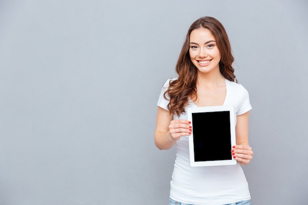 Cheerful lovely young woman standing and holding blank screen tablet