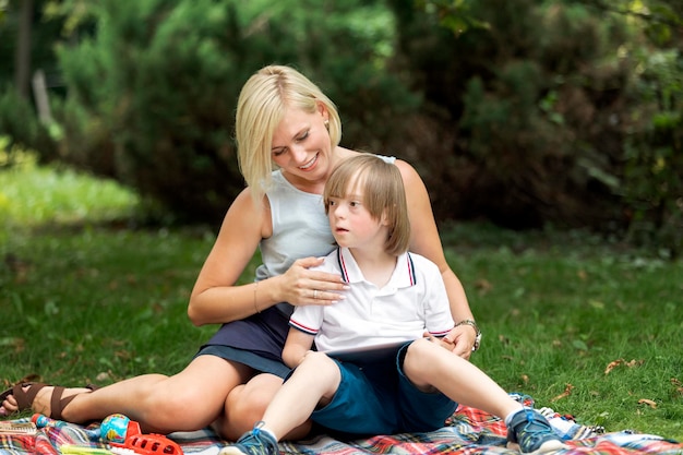 Cheerful loving mother hugging her Down syndrome son sitting on a rug on a lawn