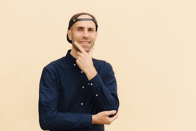 Cheerful male adult holds chin and looks thoughtfully into distance happy to hear positive news wears fashionable shirt poses against beige background with copy space for your advertisement