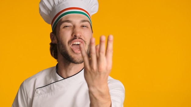 화려한 배경에 격리된 유니폼을 입고 맛있는 제스처를 보여주는 쾌활한 남성 요리사 요리사 모자를 쓰고 장난을 치는 매력적인 남자