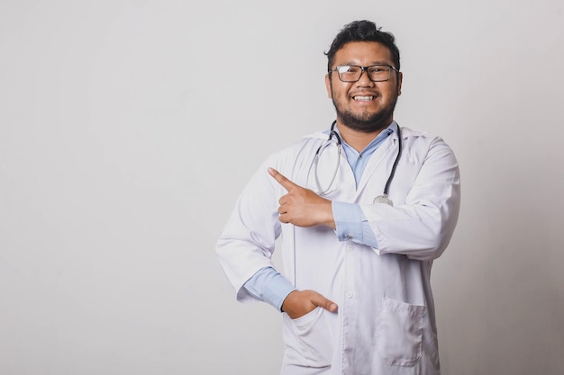 Cheerful male doctor with sideways pointing gesture at copy space isolated on white background