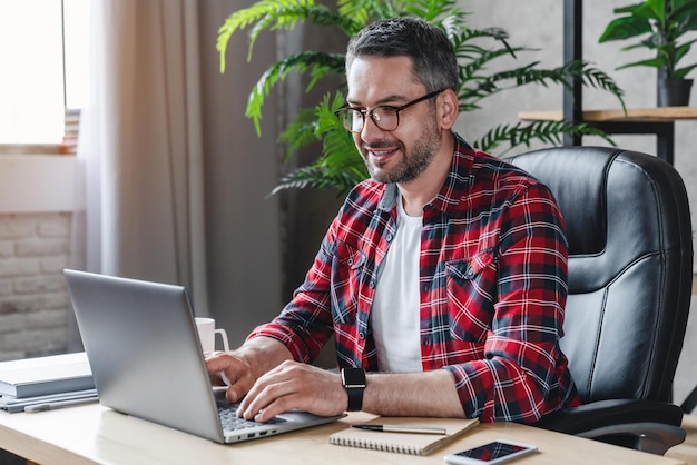 Cheerful male freelancer in eyeglasses sitting at work desktop while using notebook