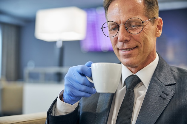 Un uomo allegro con occhiali e guanti in lattice si sta godendo il caffè mentre è seduto nella lounge dell'aeroporto