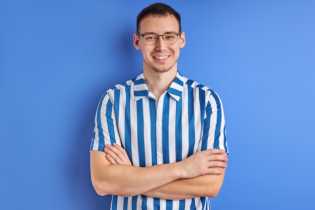 Cheerful Male in Striped Shirt In Eyeglasses posing With Arms Folded And Smiling, Isolated On Blue Studio Background