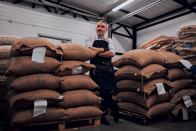 Cheerful man in apron posing standing confidently with arms crossed near coffee bags Full length