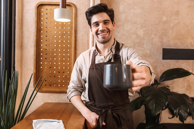 Foto uomo allegro barista che indossa un grembiule in piedi al bar, mostrando la tazza di caffè