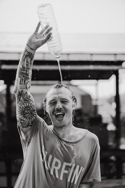 A cheerful man in a bright t-shirt with a skateboard in a skatepark pours water from a bottle on his head.