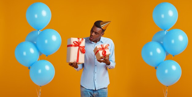 Cheerful man in cap holding two gift boxes with red ribbons, yellow background. Smiling male person got a surprise, event or birthday celebration, balloons decoration