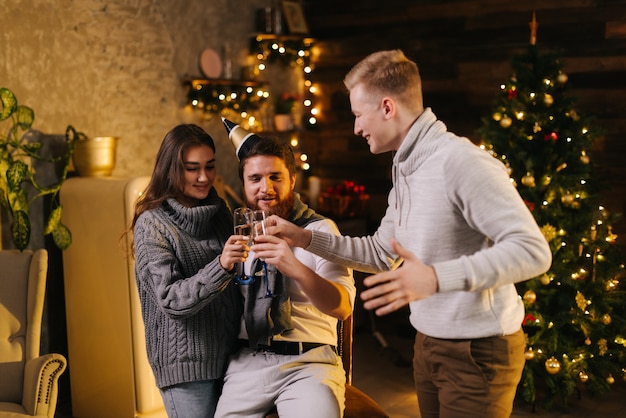 Cheerful man clinking champagne glasses with couple in celebrating new years eve party in festively decorated house. Christmas tree with garland and festive illumination in background.