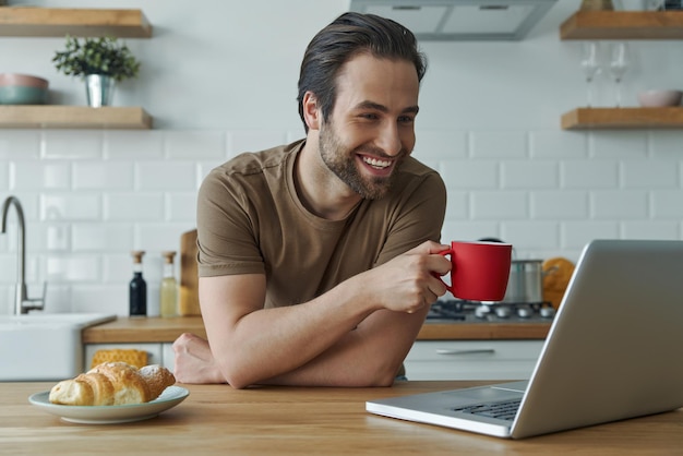 キッチンアイランドでラップトップで働いている間朝のコーヒーを楽しんでいる陽気な男性