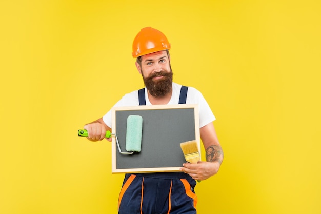 Cheerful man in helmet and work clothes hold paint roller brush and blackboard with copy space on yellow background