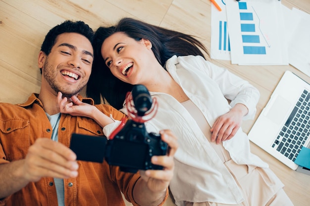Cheerful man holding a camera and recording himself while lying on the floor with his happy girlfriend