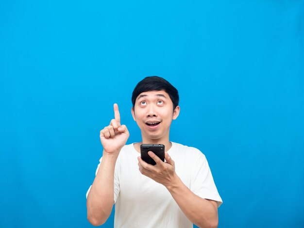 Cheerful man holding cellphone and point finger up at copy space blue background